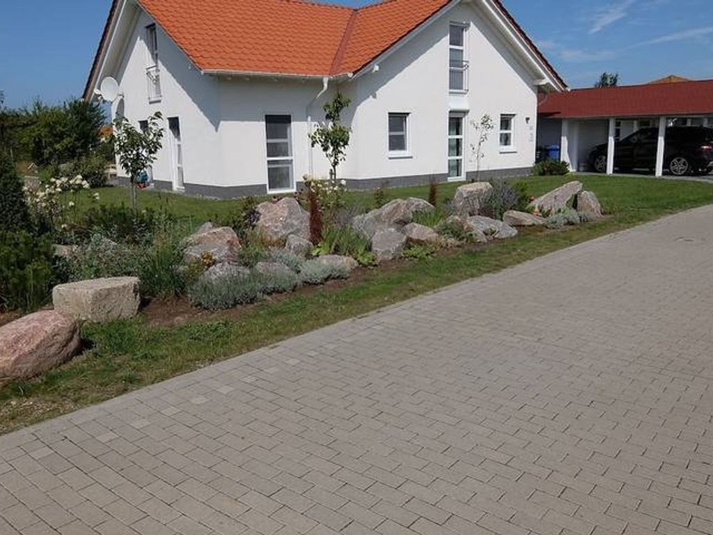 Ein zweigeschossiges Haus mit rotem Dach, einem gepflasterten Auffahrt und einem Garten mit großen Steinen und Sträuchern.