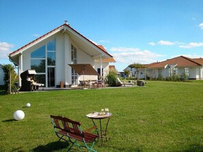 Ein Wohnhaus mit großem Vorgarten, einer Terrasse und Gartenmöbeln, vor blauem Himmel.