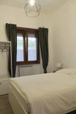 Une chambre à coucher comprenant un lit, un radiateur, une fenêtre avec des rideaux, un porte-manteau et un luminaire suspendu.