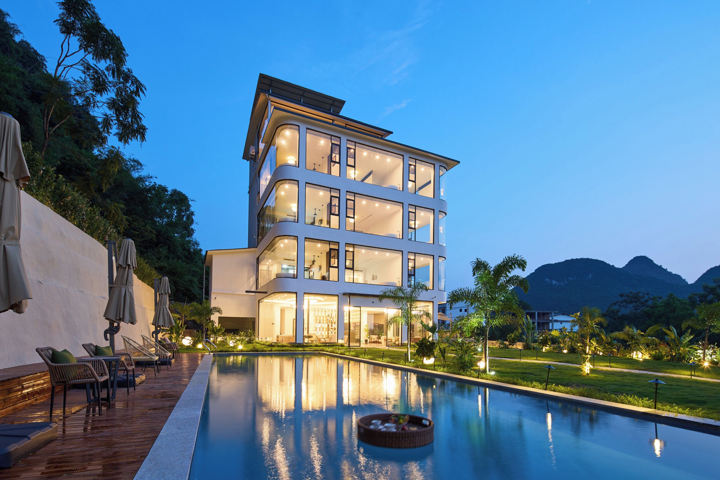 Un hotel de varios pisos con piscina, mobiliario exterior y montañas al fondo.