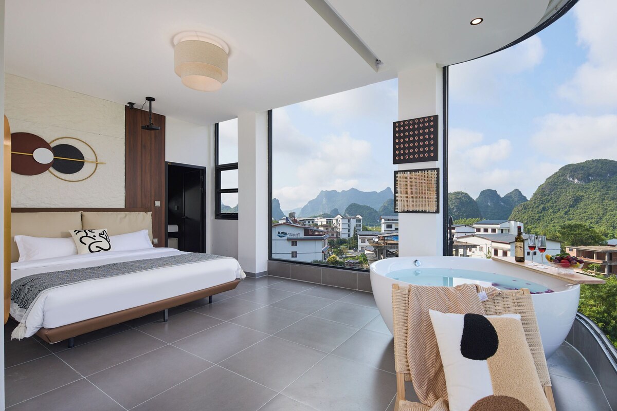 A modern hotel room with a large bed, a bathtub, and a scenic mountain view.