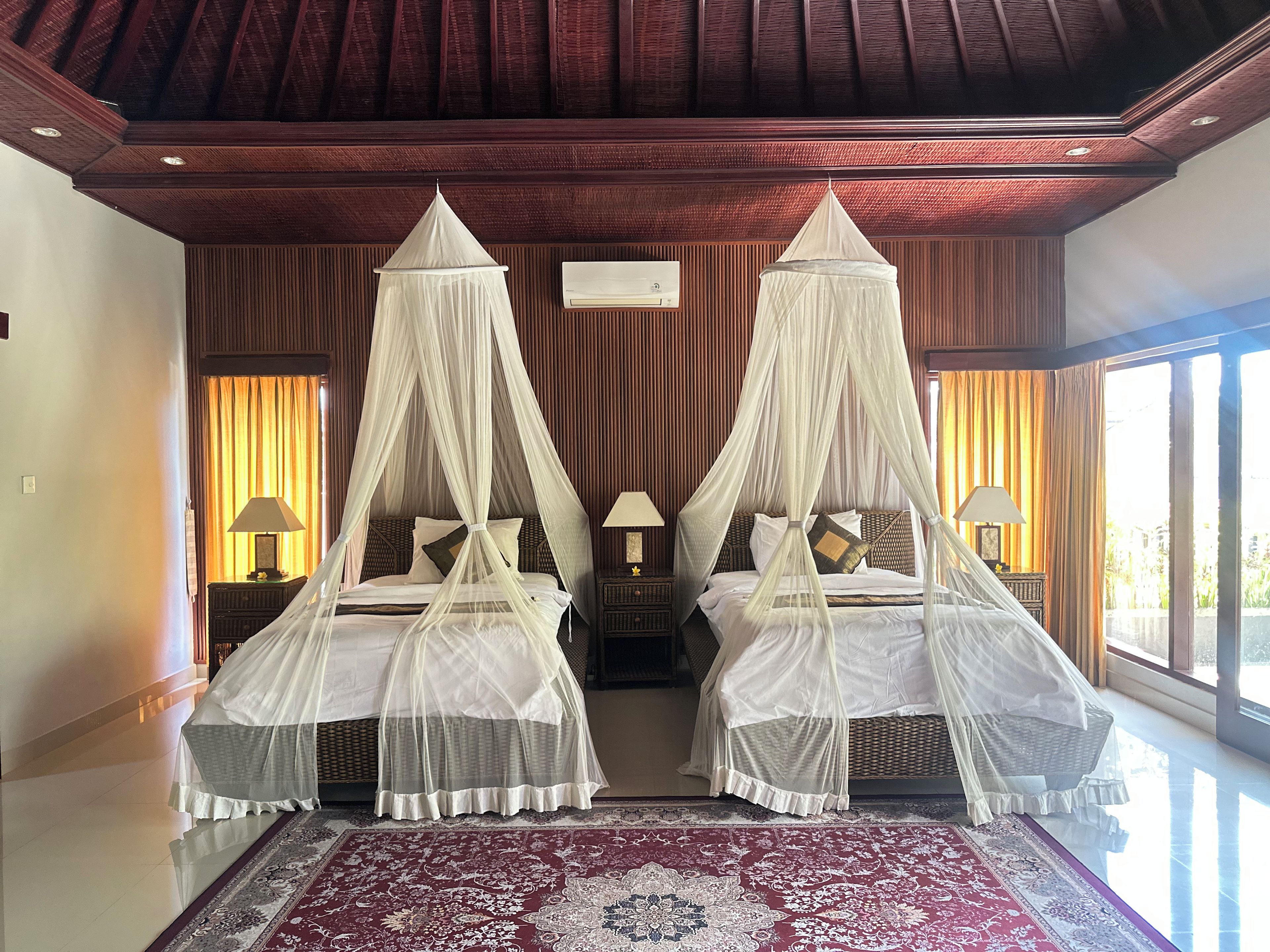 A double bed with a canopy, a nightstand with a lamp, and a large window with curtains.