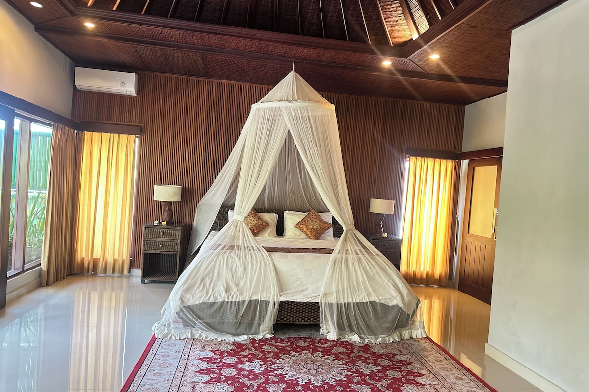 A room with a four-poster bed, a red patterned carpet, wooden paneling, and a large window with curtains.