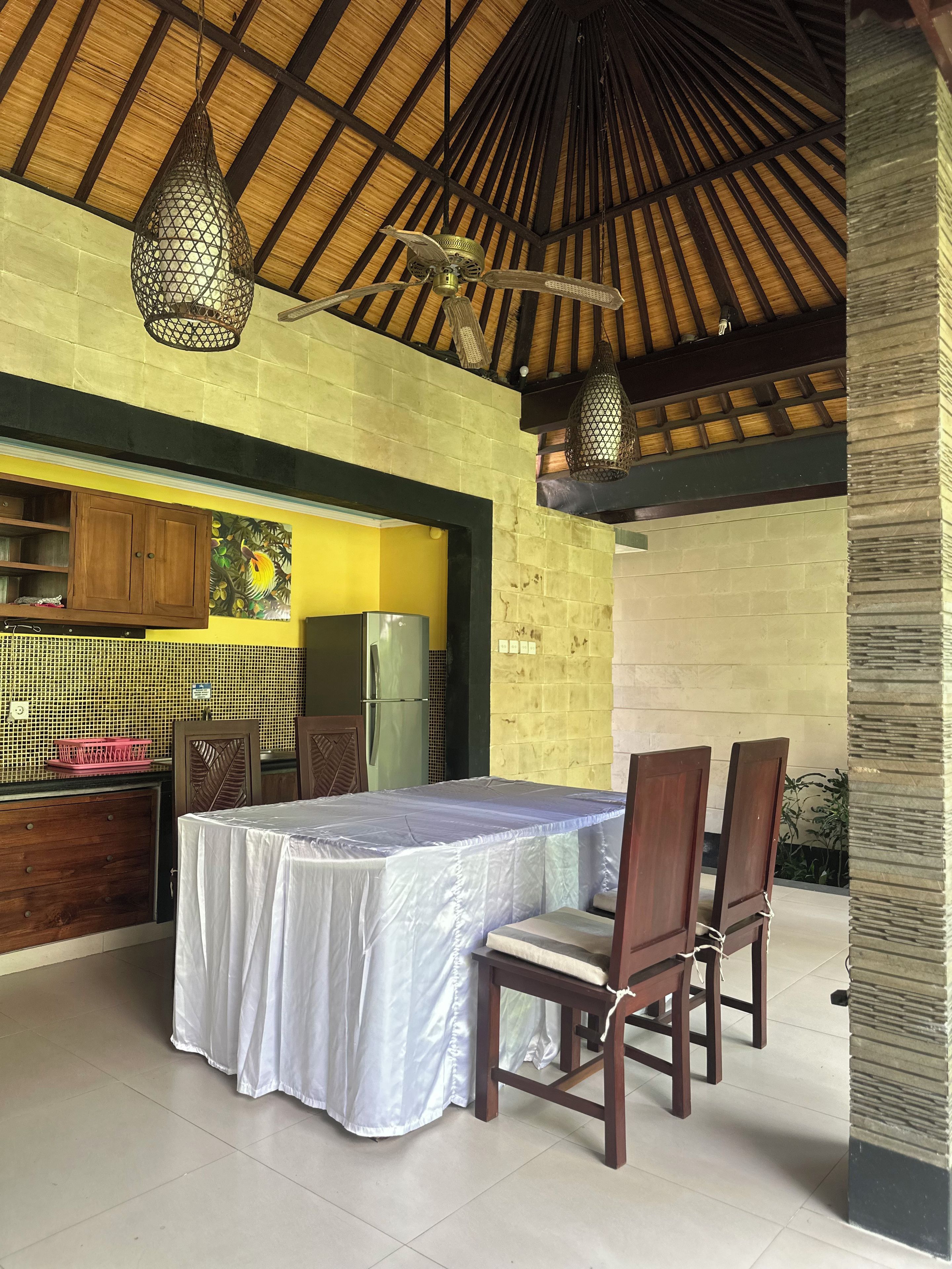 An outdoor dining area with a table and chairs, a ceiling fan, and a kitchen area with a refrigerator.