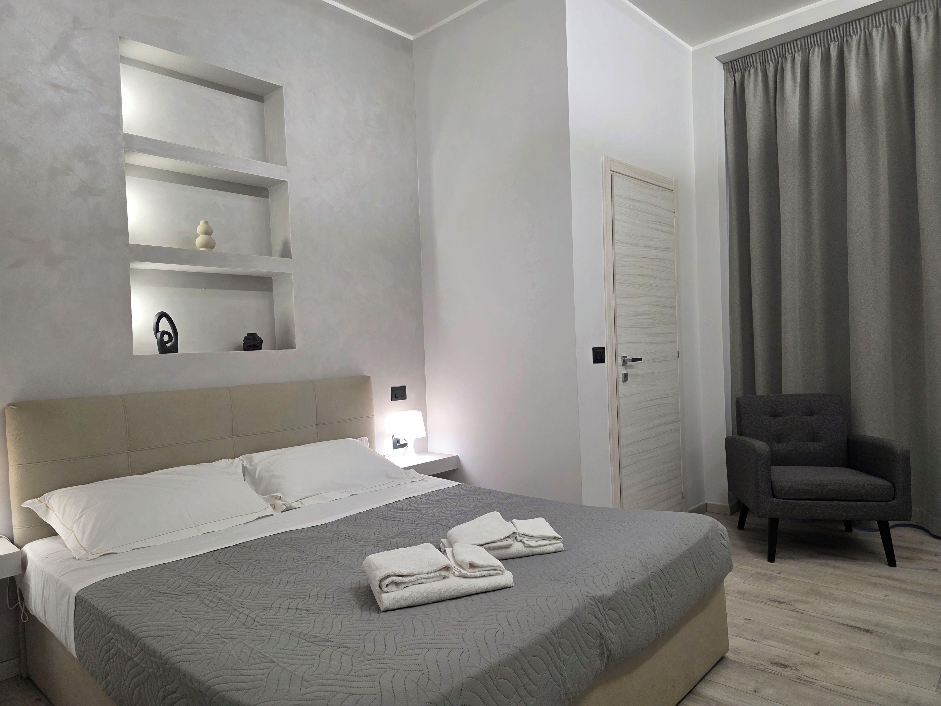 Una habitación de hotel con cama, mesitas de noche, una silla, una puerta y una ventana con cortinas.