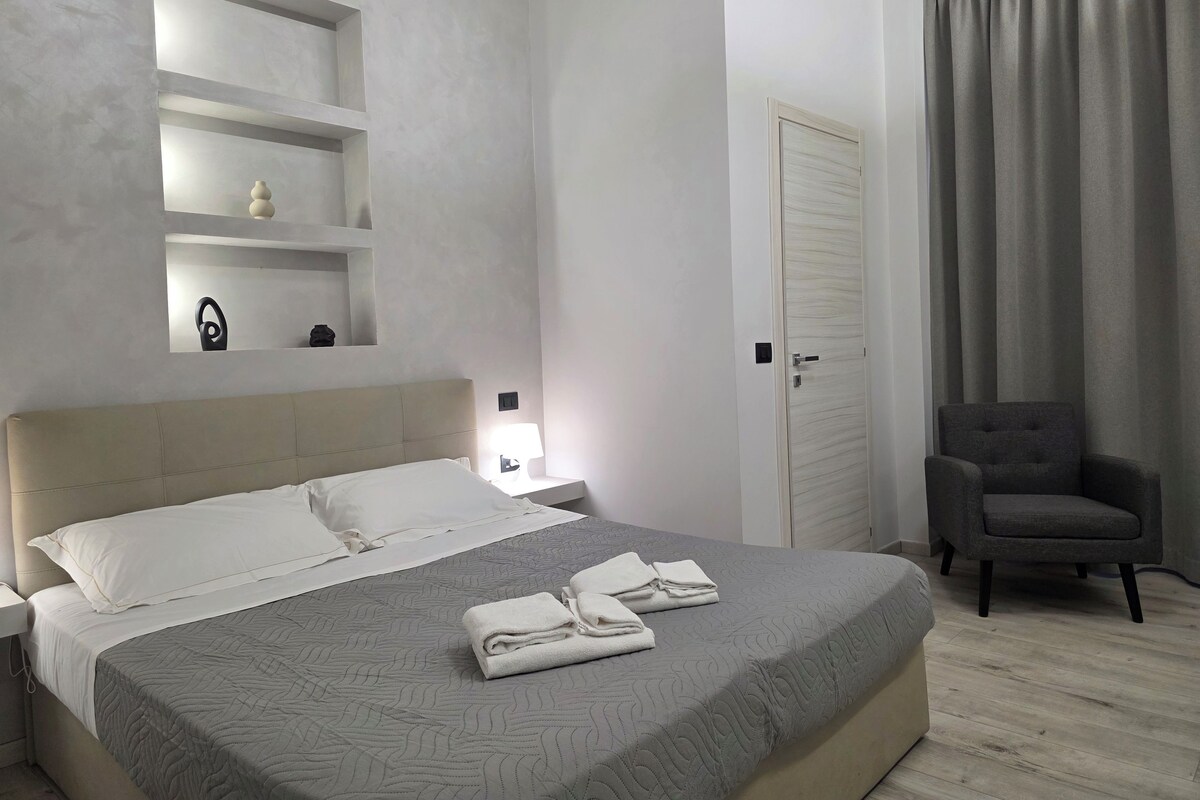 Una habitación de hotel con cama, mesitas de noche, una silla, una puerta y una ventana con cortinas.