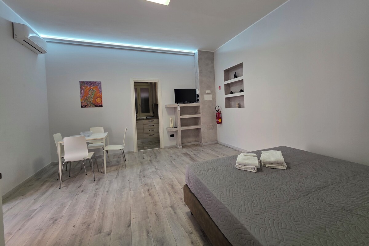 Un dormitorio moderno con cama, escritorio con sillas, televisor y estante con objetos decorativos.