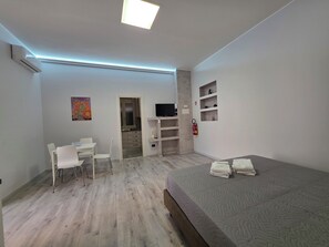 Un dormitorio moderno con cama, escritorio con sillas, televisor y estante con objetos decorativos.