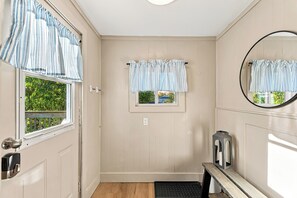 Een badkamer met een houten vloer, een witte deur met een zilveren handvat, een raam met uitzicht op groen, een klein raam met uitzicht op een huis, en een spiegel aan de muur.