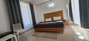 A modern bedroom with a wooden headboard, a bed with a dark blue bedspread, white pillows, a white chair, and a small table. - Solang Valley Homestay (Manali)