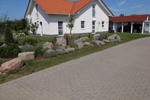 Et hvidt hus med rødt tag, en belagt indkørsel og en have med store sten og buske.