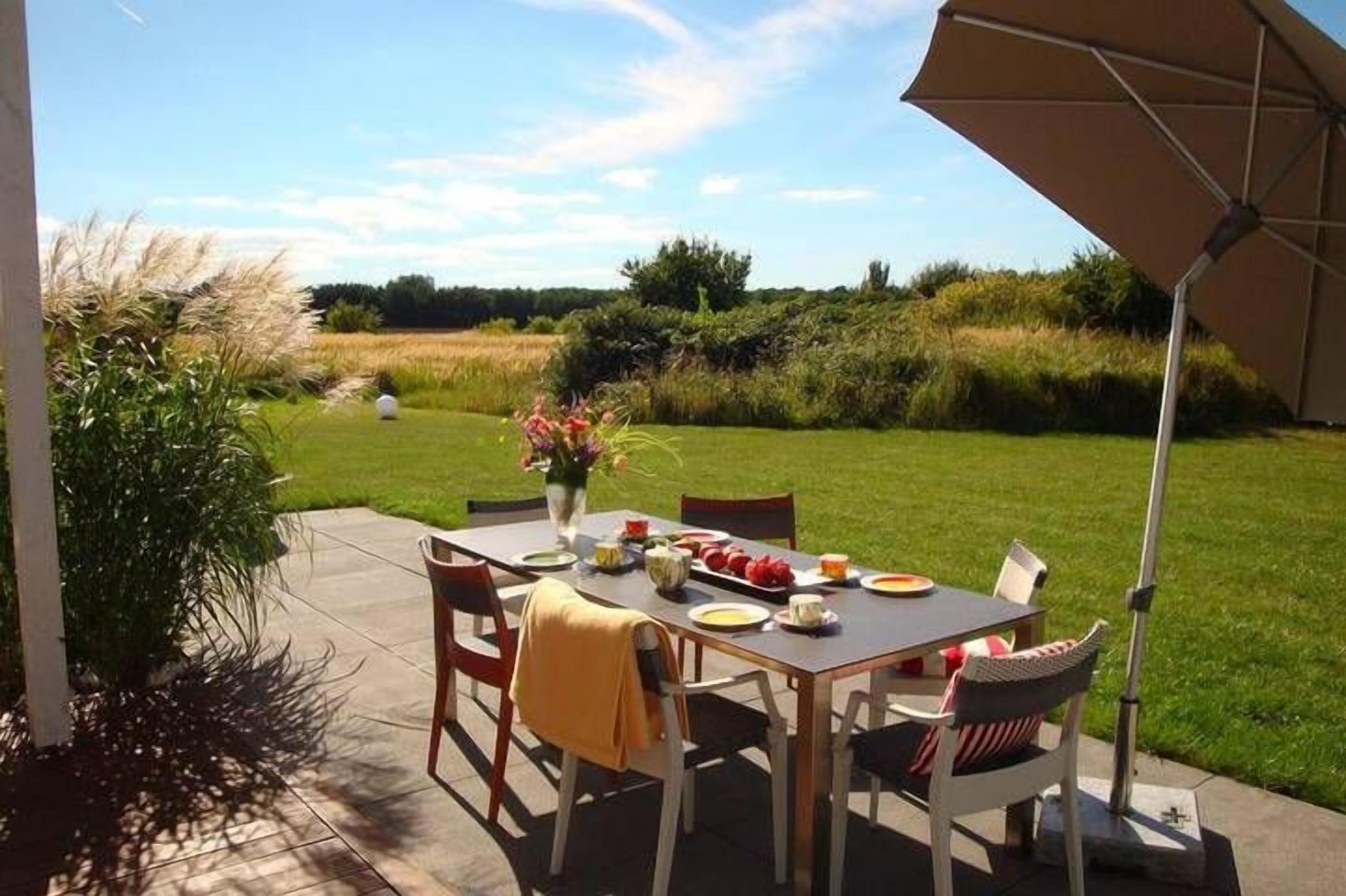 En udendørs terrasse med et bord dækket til to personer, omgivet af stole, en parasol og en vase med blomster.