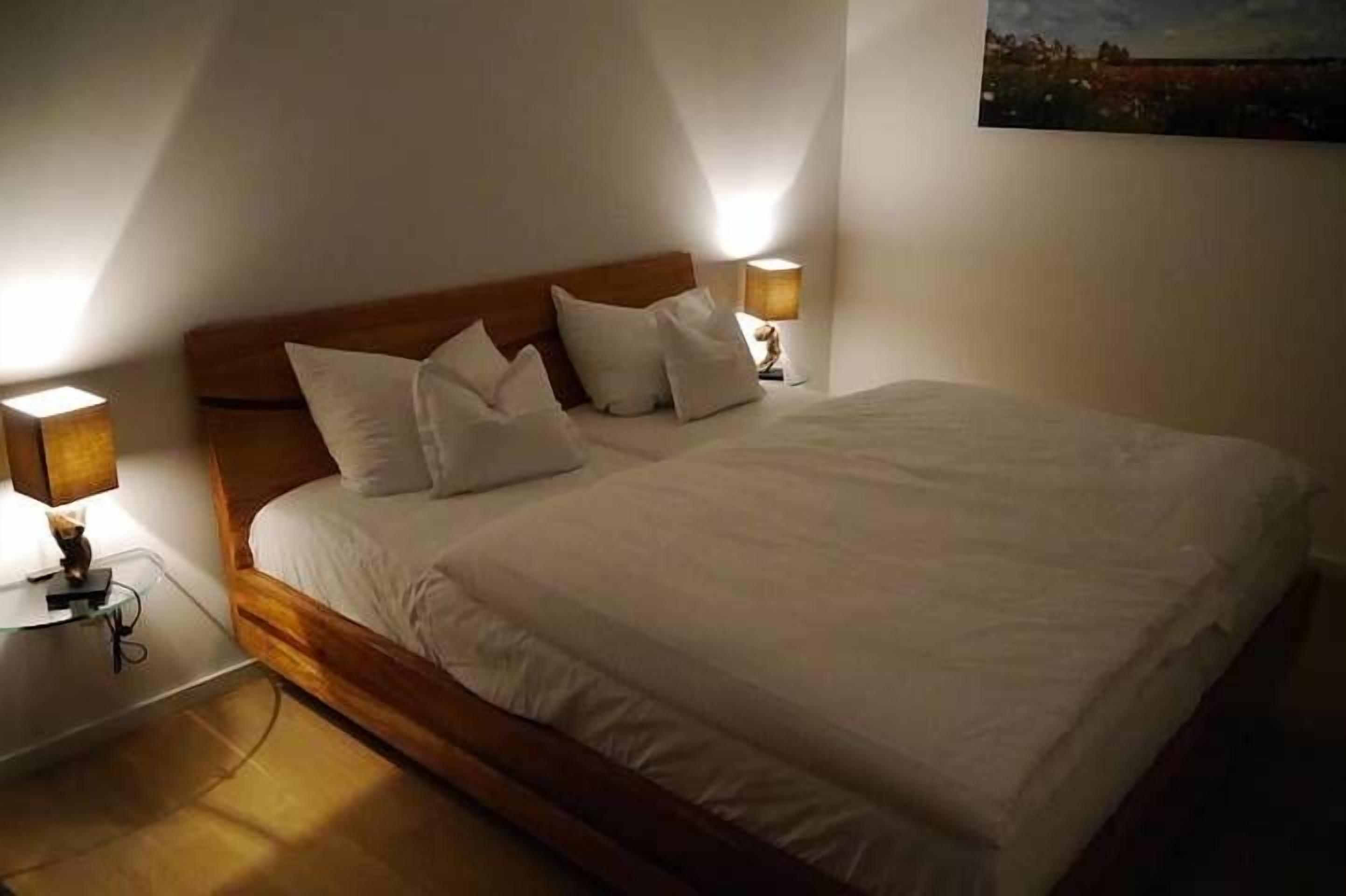 En pænt redt seng med hvide sengetøj og puder, to sengeborde med lamper, og et maleri på væggen.