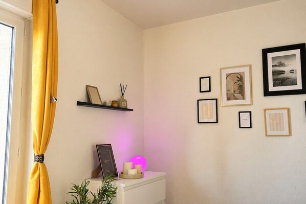 A room with a white bedside table, a shelf with framed pictures, and a yellow curtain. - New Listing (Lucé)