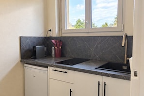 A kitchen with a built-in dishwasher, a black countertop, a window above the sink, and a view of greenery outside. - New Listing (Lucé)