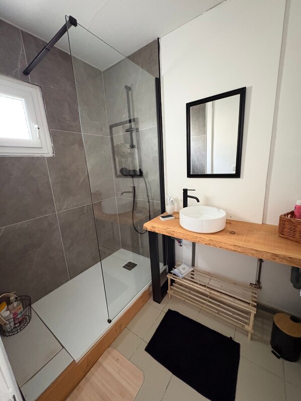 Un baño moderno con ducha envuelta en vidrio, un lavabo blanco y un mueble de tocador de madera.