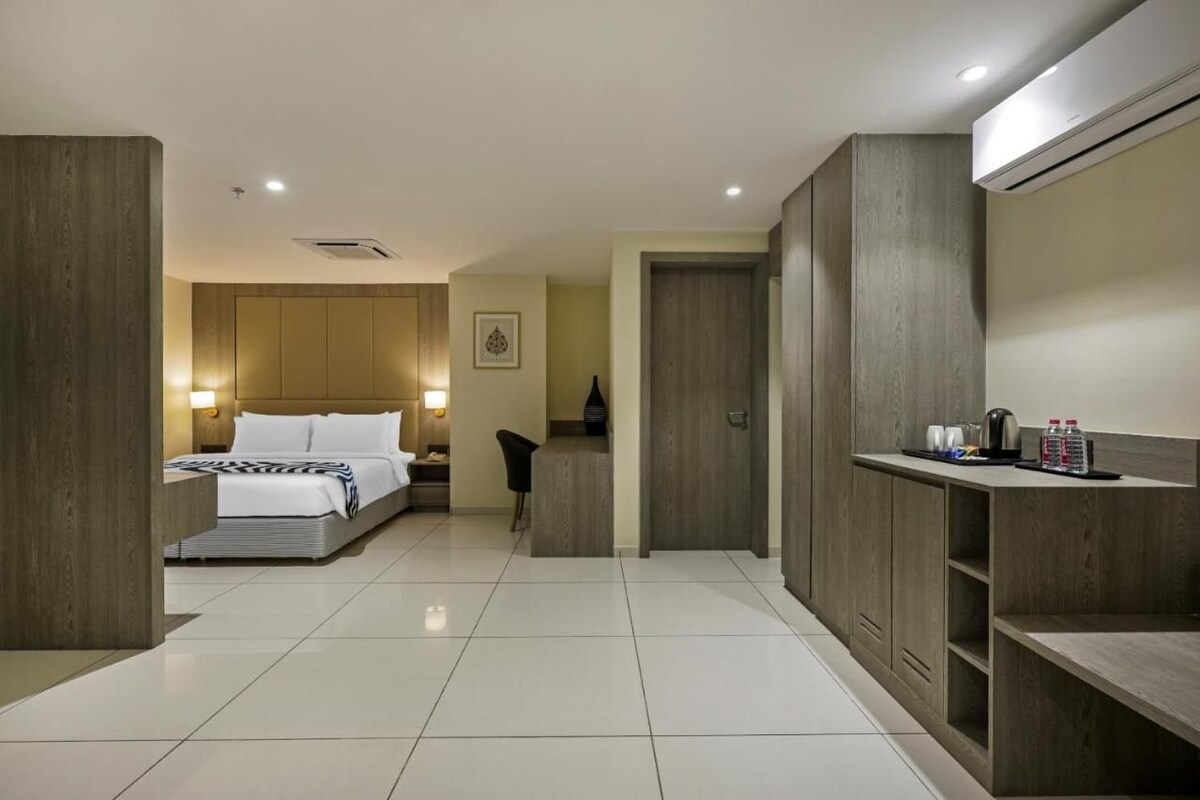 A hotel room with a bed, bedside table, lamp, wardrobe, and a small kitchenette area.