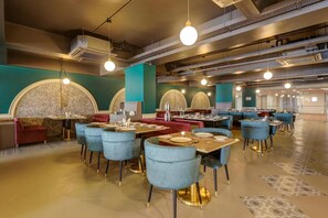 A spacious indoor dining area with round tables and chairs, a central seating area with a red sofa, and a patterned wall. - POP 84978J Sunday Vadodara Palace Hotel (Vadodara)