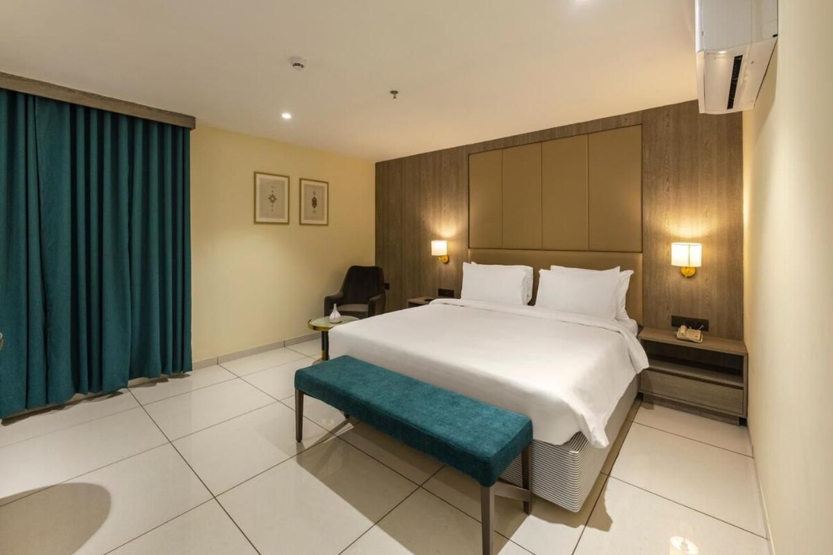 A hotel room with a large bed, bedside tables, a bench, and a wall-mounted air conditioner.