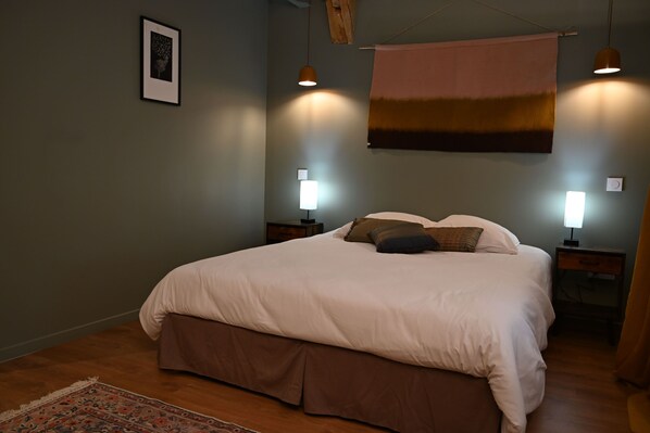 Une chambre à coucher avec un lit, deux tables de chevet, une commode, une lampe suspendue, un tableau encadré au mur et un tapis à motifs.