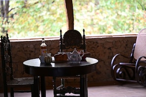 Une table ronde, sur laquelle sont posés une bouteille, une théière et du pain, est placée devant un mur de pierre, avec vue sur la verdure à l'extérieur.