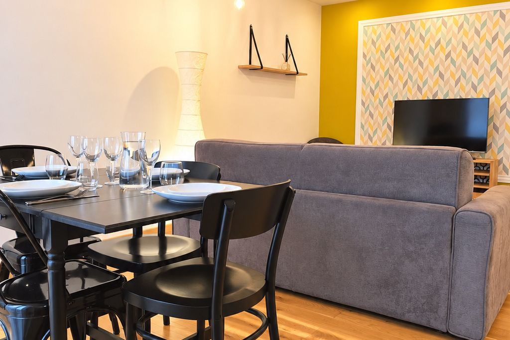 Een eetgedeelte met een tafel voor vier personen, een grijze bank en een televisie aan de muur.