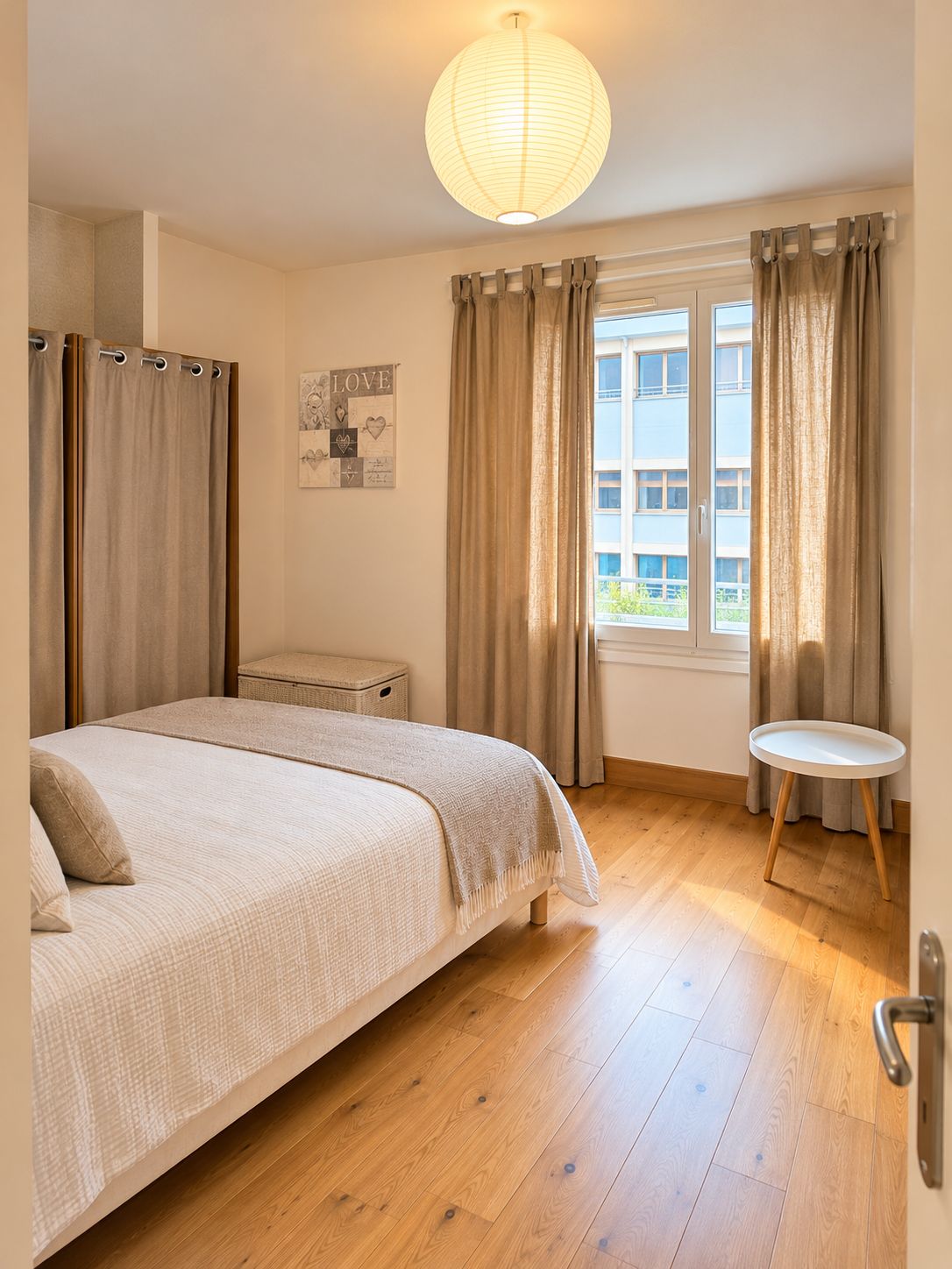 Een slaapkamer met een bed, een klein tafeltje, een raam met gordijnen en een wandlamp.