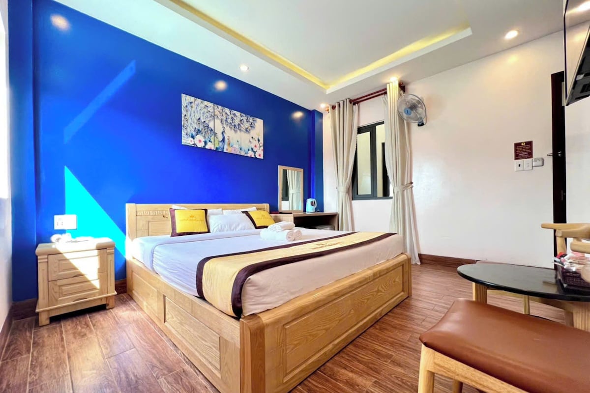 Een hotelkamer met een groot bed, een houten hoofdeinde, een nachtkastje met een lamp, een klein tafeltje met een stoel en een raam met gordijnen.