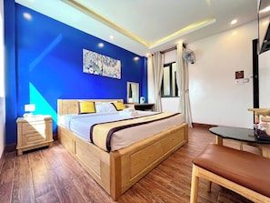 Een hotelkamer met een groot bed, een houten hoofdeinde, een nachtkastje met een lamp, een klein tafeltje met een stoel en een raam met gordijnen.