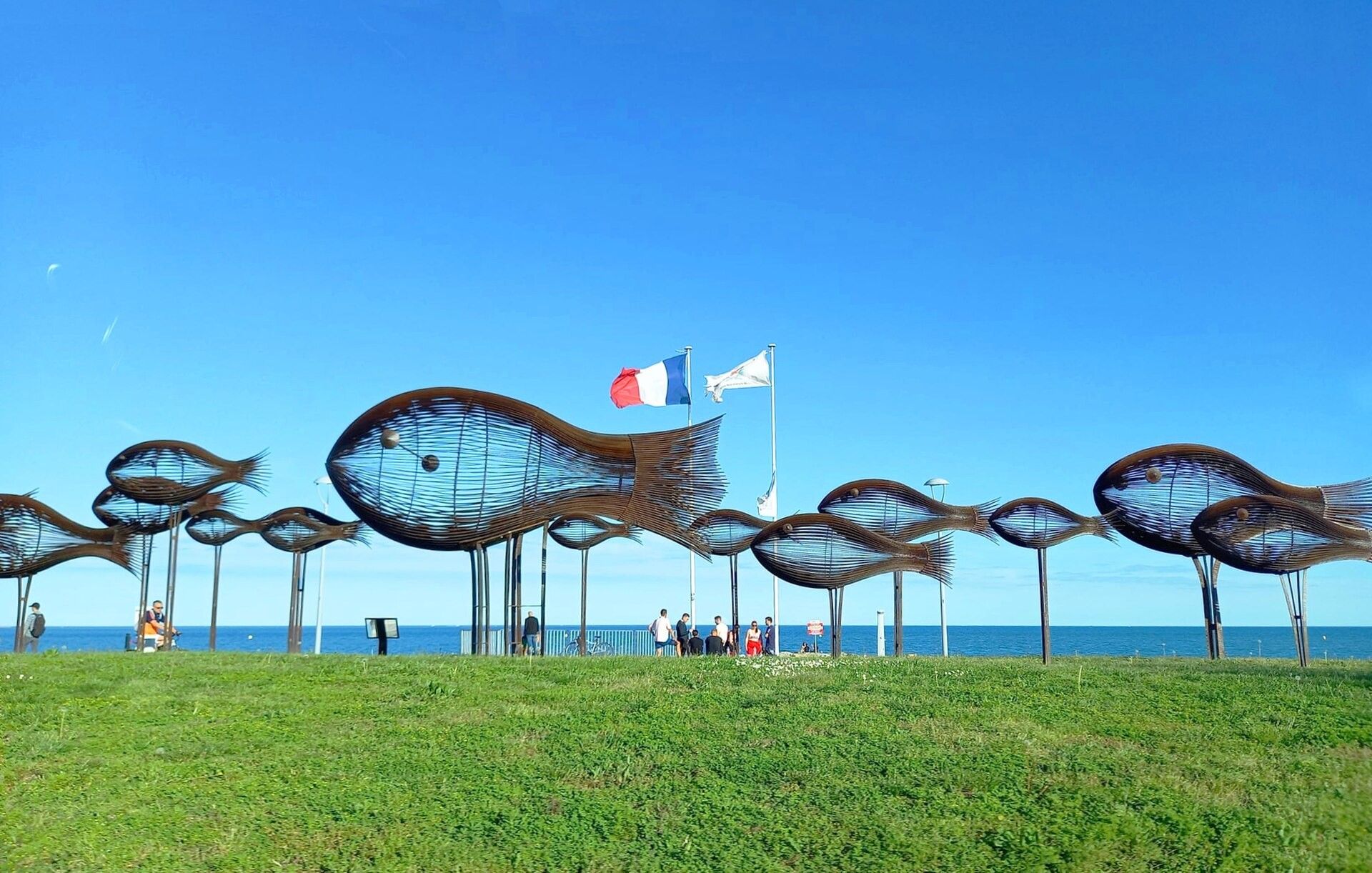 A coastal area with a series of metal sculptures resembling fish, a flagpole with a flag, and a clear view of the sea.
