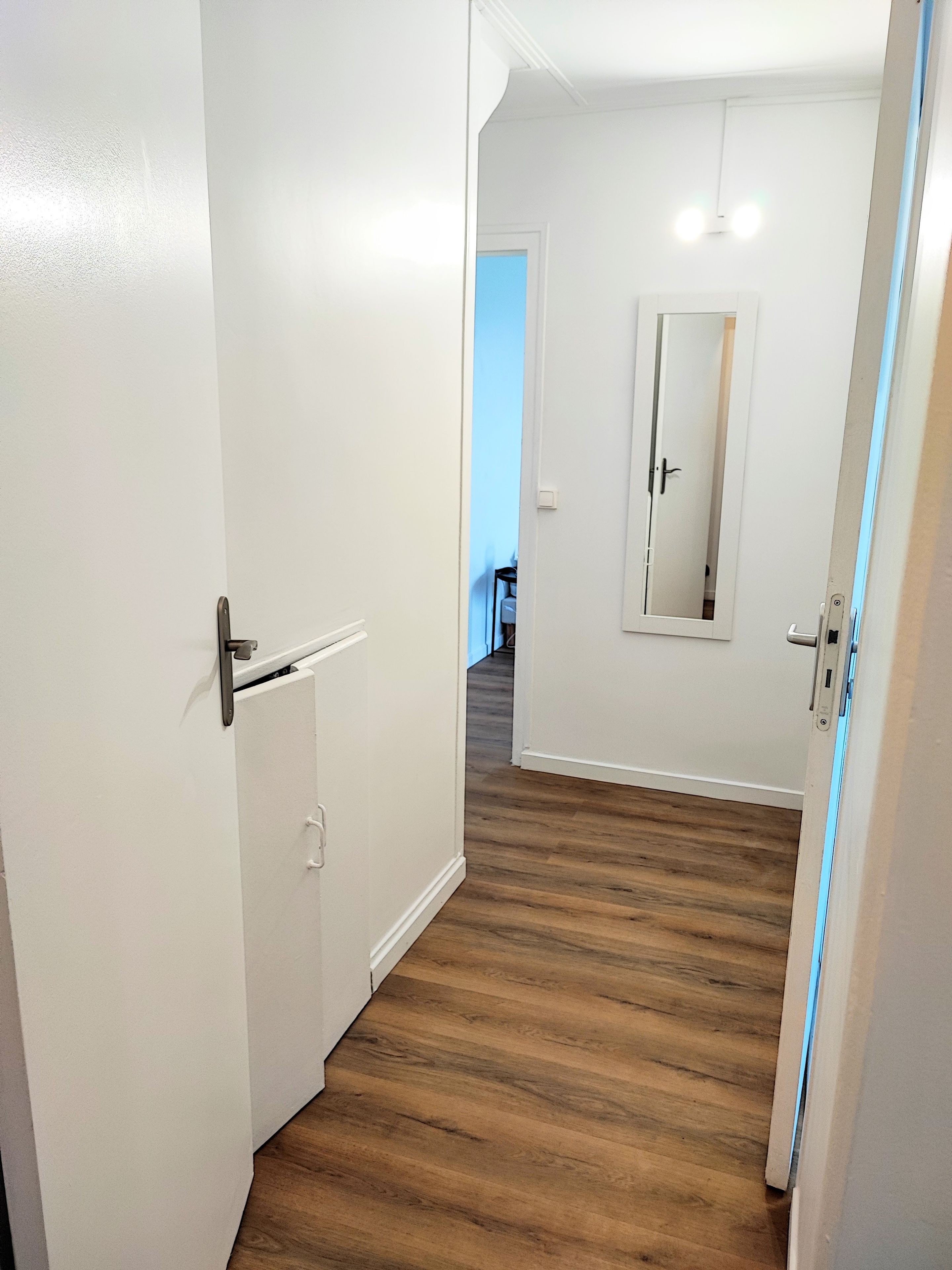 A hallway with wooden flooring and white walls, leading to a room with a door and a mirror on the wall.