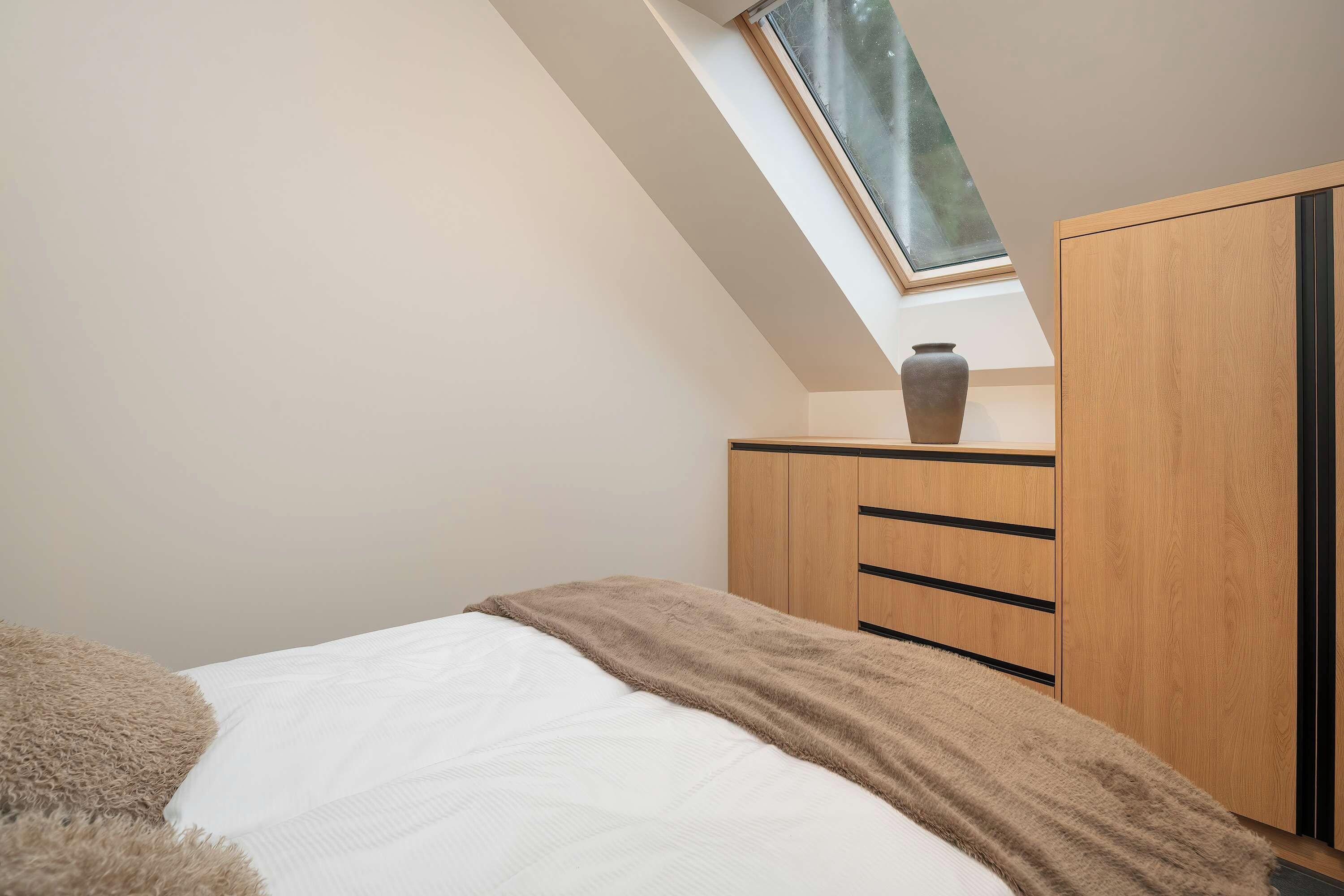 Una camera da letto con soffitto spiovente, una finestra a lucernario, un letto con materasso bianco e coperta marrone, e un armadio in legno con maniglie nere.