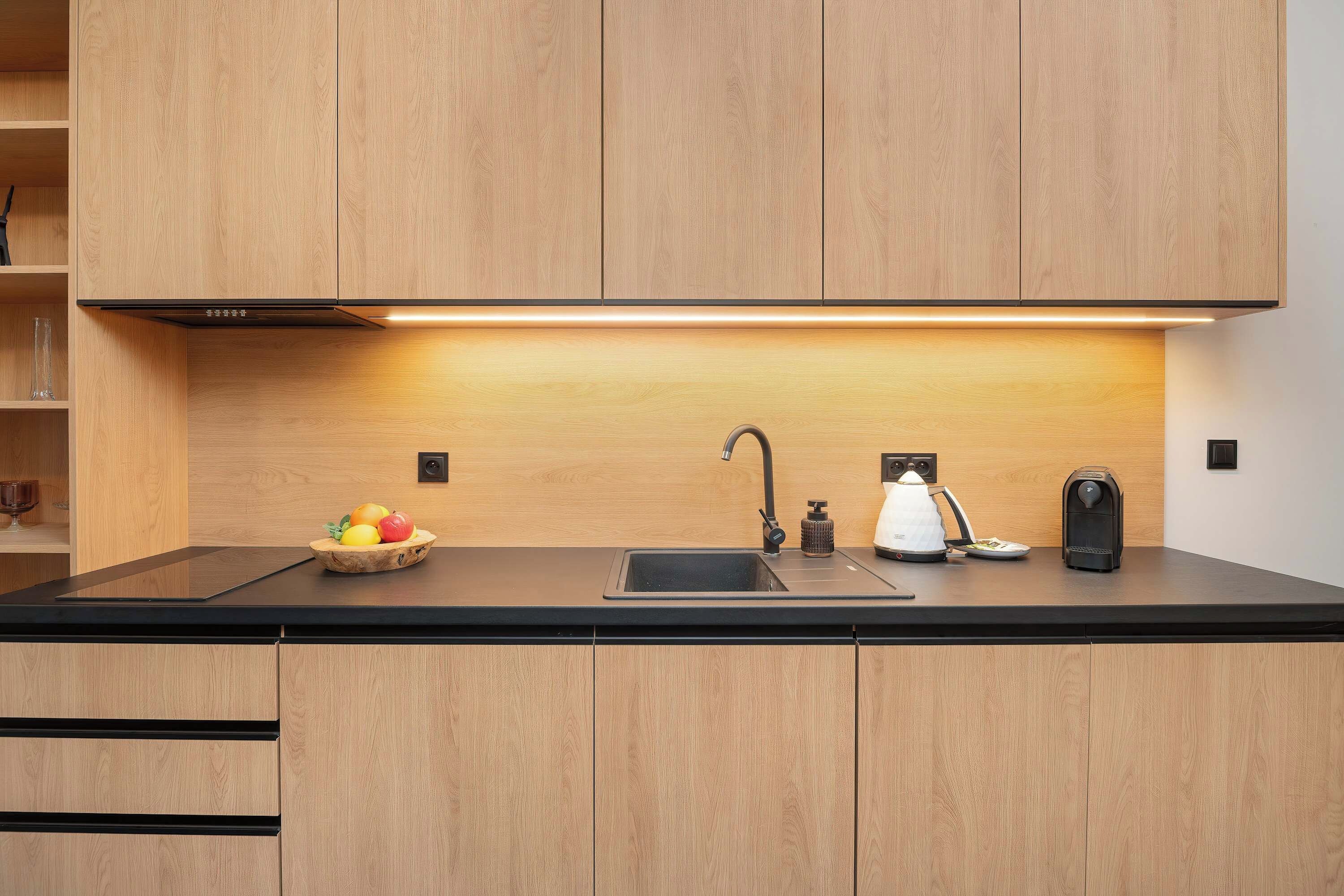 Una cucina moderna con un piano di lavoro nero, mobili in legno, un lavello incassato, un bollitore, una macchina da caffè e una ciotola di frutta.