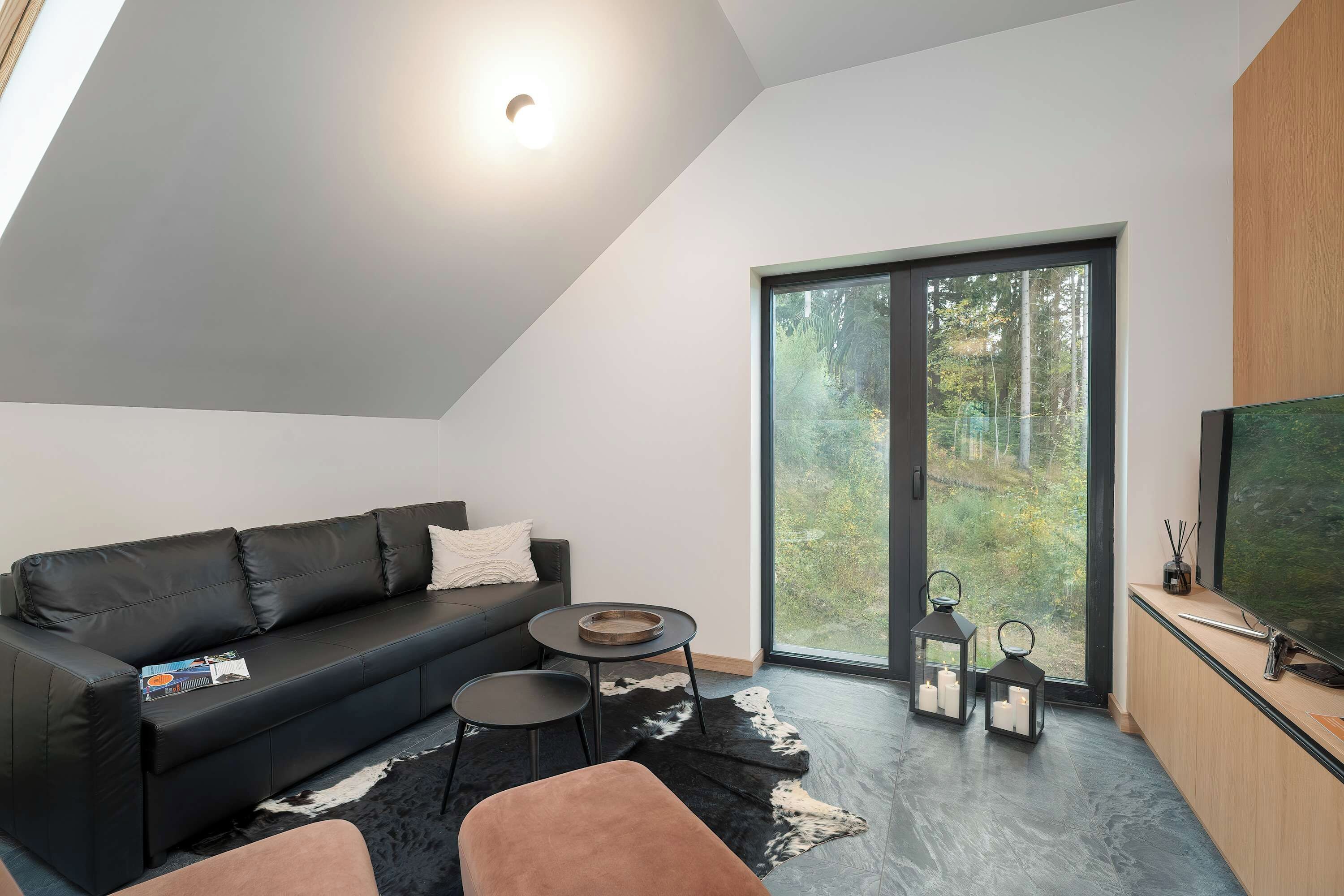 A modern living space with a black leather sofa, a round coffee table, a black and white rug, and a large sliding glass door leading to a forested area.