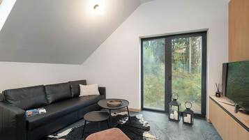 A modern living space with a black leather sofa, a round coffee table, a black and white rug, and a large sliding glass door leading to a forested area.