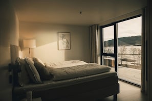 A bedroom with a bed, a bedside lamp, a framed picture, and a large window with a view of a landscape.