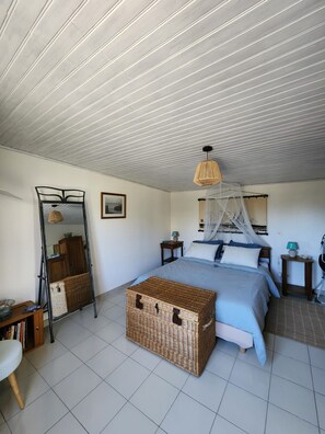 Ein Schlafzimmer mit einem Bett, einem Nachttisch, einem Beistelltisch, einer Korbschachtel, einem Spiegel und einer Pendelleuchte.