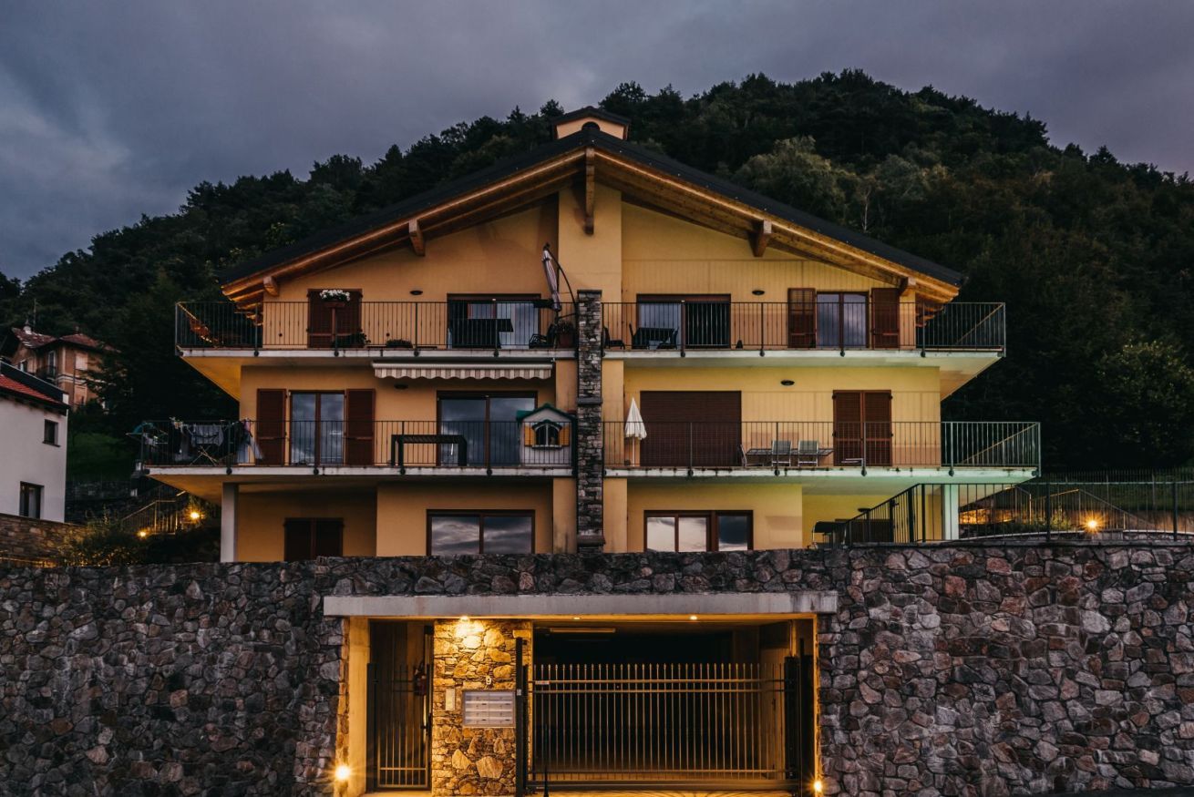 Un edificio a due piani con balconi, un muro di pietra in primo piano e una montagna sullo sfondo.