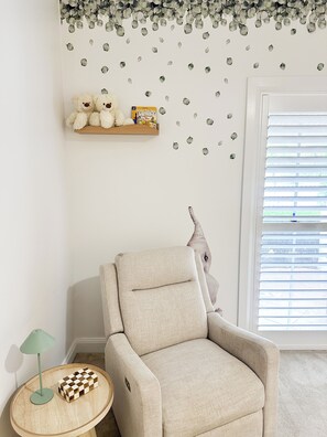 A room with a beige armchair, a wooden side table with a lamp, a shelf with books and a teddy bear, and a wall with leaf decals.