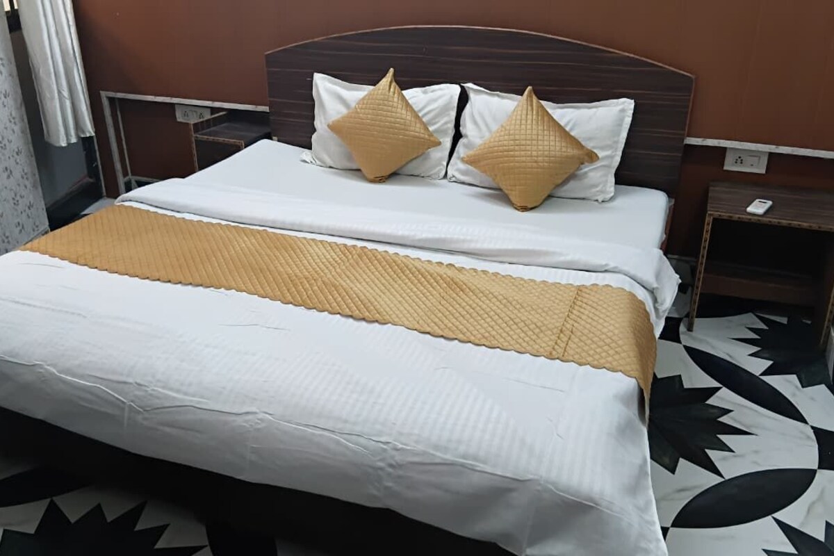 Een tweepersoonsbed met een wit en beige sprei, twee decoratieve kussens, een nachtkastje met een telefoon, en een vloer met een patroon.