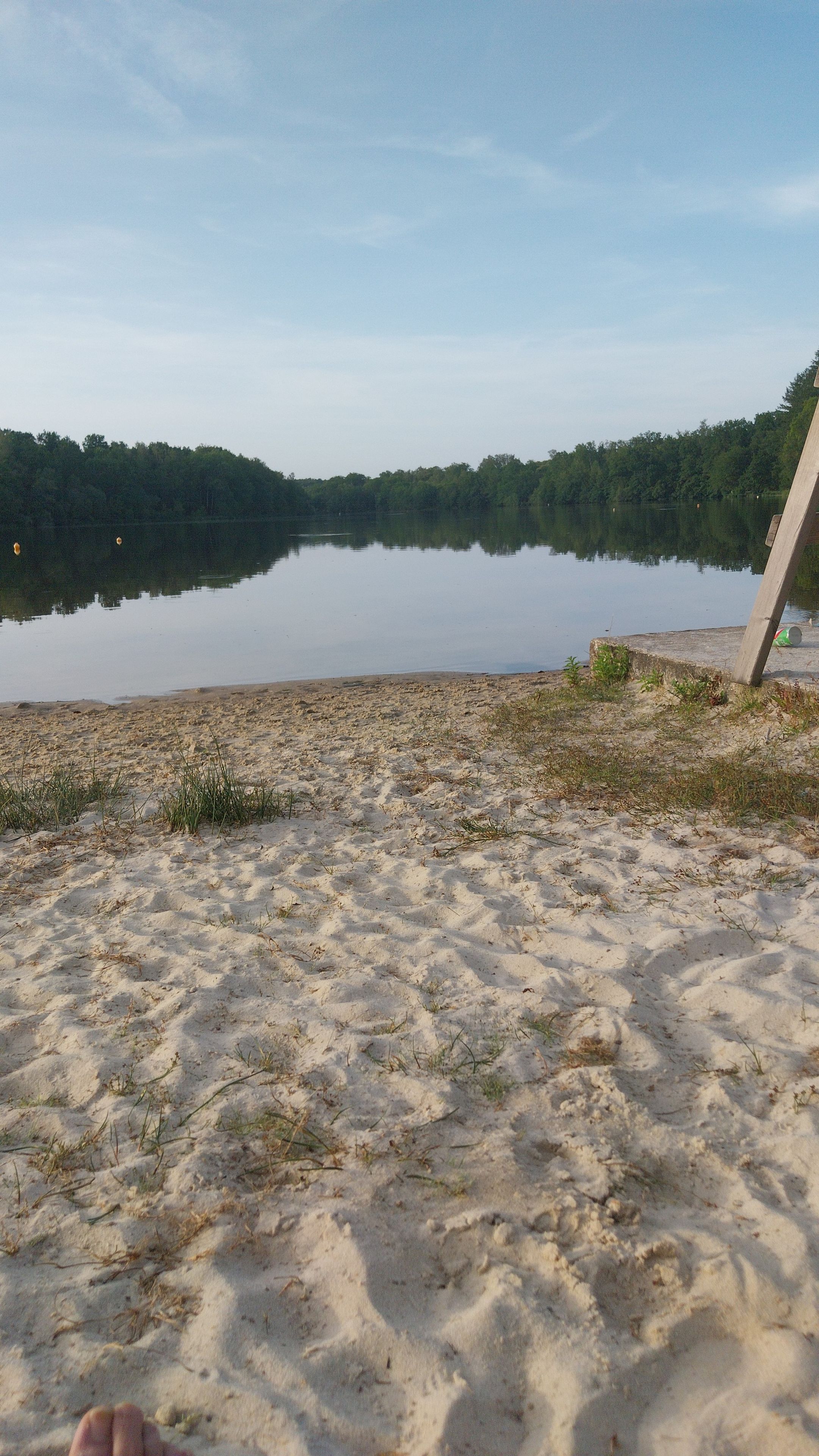Une plage de sable bordée d’un lac aux eaux calmes, d’une bordure en béton et d’une planche de bois tombée à terre.
