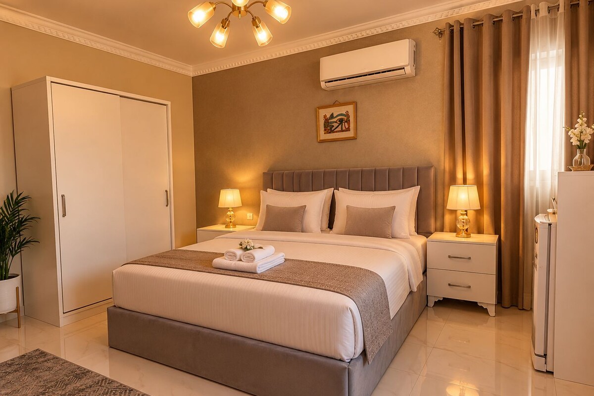 A hotel room with a bed, bedside tables, a lamp, an air conditioner, a potted plant, and a door.