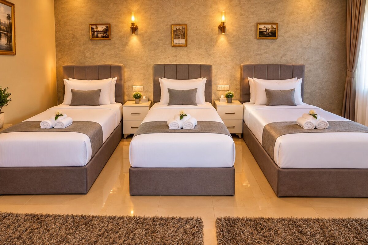 A hotel room with three beds arranged in a row, each with a headboard, bedside tables, and towels.