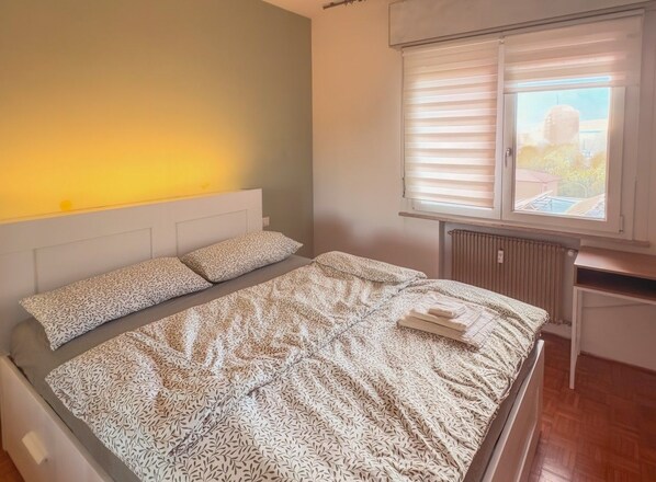 A bedroom with a bed, headboard, window with blinds, radiator, and a small table. - Pordenone House - Near the Historic Center (Pordenone)