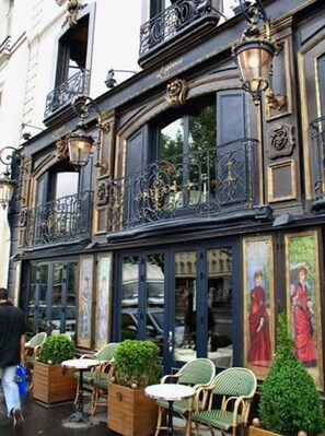 An outdoor seating area with tables and chairs, a building facade with ornate ironwork, and a street view. - Elegant Seine-Side Apartment Near Notre-Dame Cathedral & the Louvre (Paris)