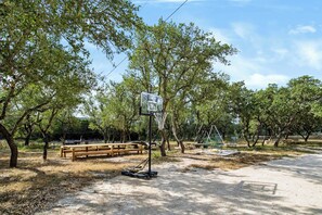 A basketball court with a wooden bench and chairs, surrounded by trees. - Mcfoy Ranch 20 Acres, Sleeps 8 (Wimberley)