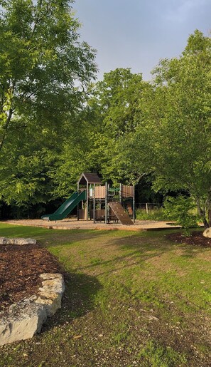 Un parque infantil con tobogán y estructuras para escalar, rodeado de árboles y césped.