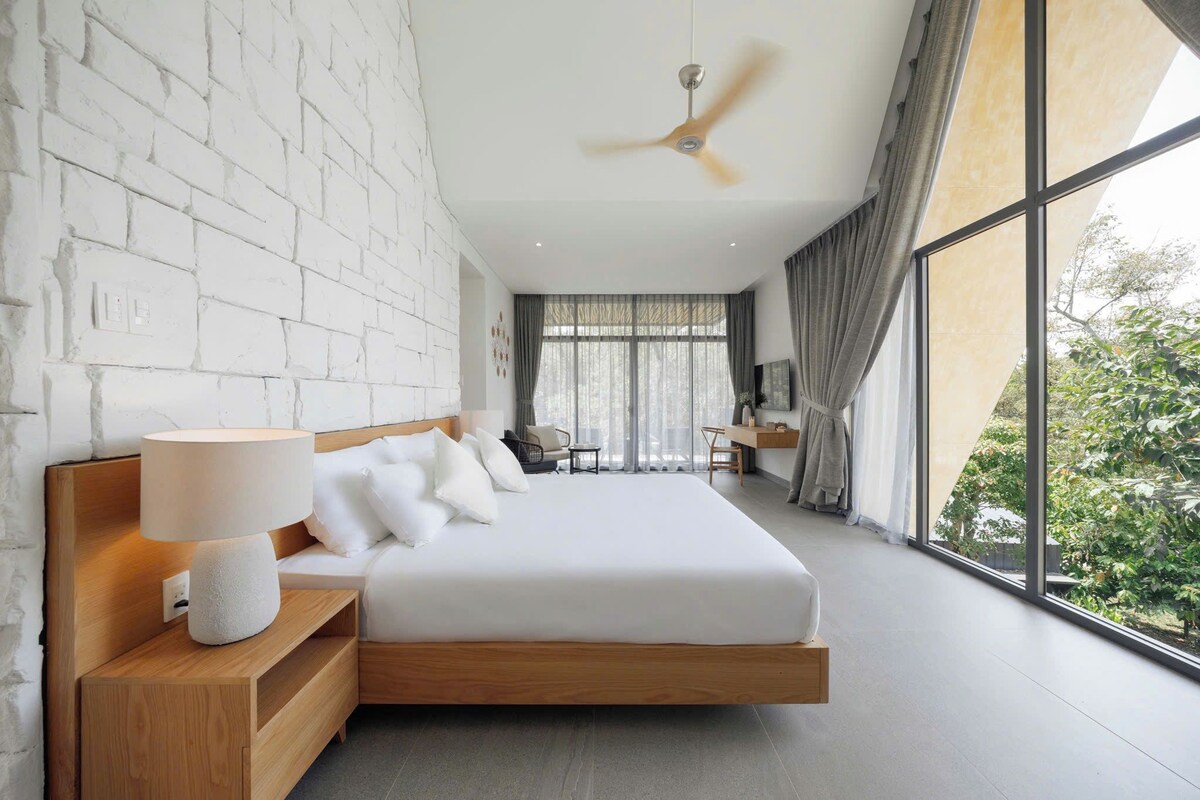 A modern bedroom with a large bed, a ceiling fan, and a view of greenery outside.