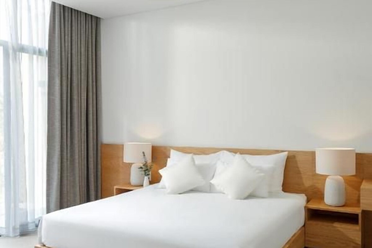 A hotel room with a large bed, wooden headboard, bedside lamps, a ceiling fan, and a view of the outdoors through the window.