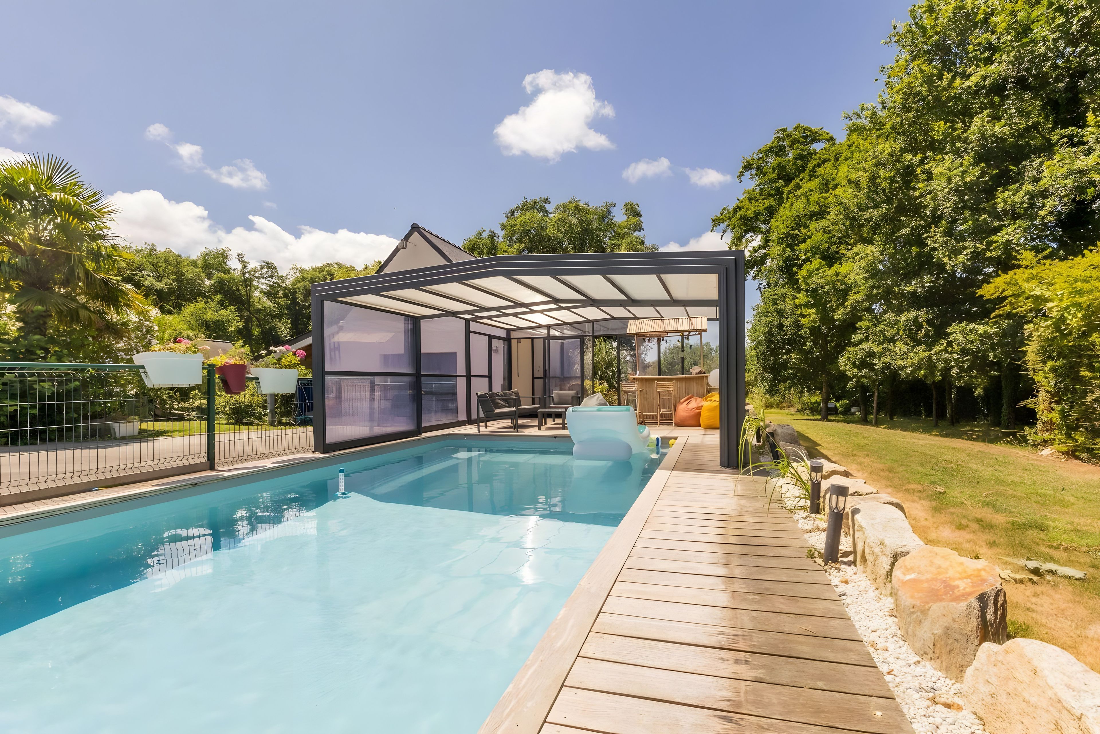 Une piscine entourée de parois en verre, dotée d’une terrasse en bois et bordée de verdure.